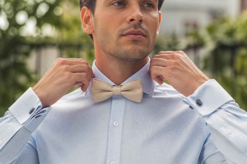 Mężczyzna w jasnoniebieskiej koszuli poprawia kołnierzyk przy beżowej muszce bow tie