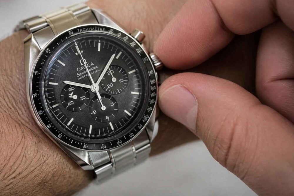 Srebrny zegarek Omega Speedmaster z czarną tarczą i chronografem na nadgarstku, regulowany dłonią.