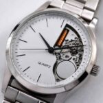 Srebrny zegarek na metalowej bransolecie z białą tarczą i odsłoniętym mechanizmem po prawej stronie